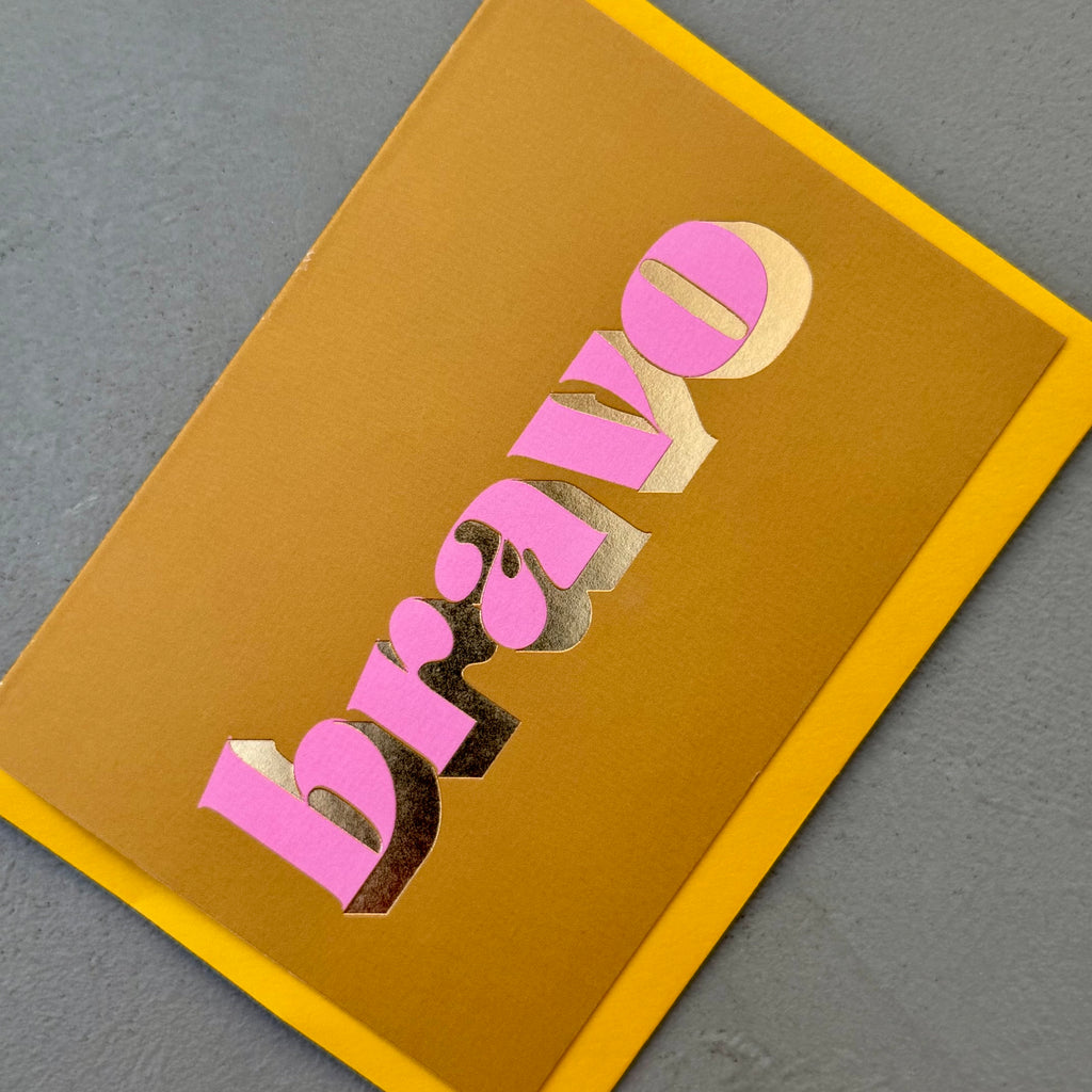Angled view of the ochre Bravo card showing the pink lettering and reflective gold foil.
