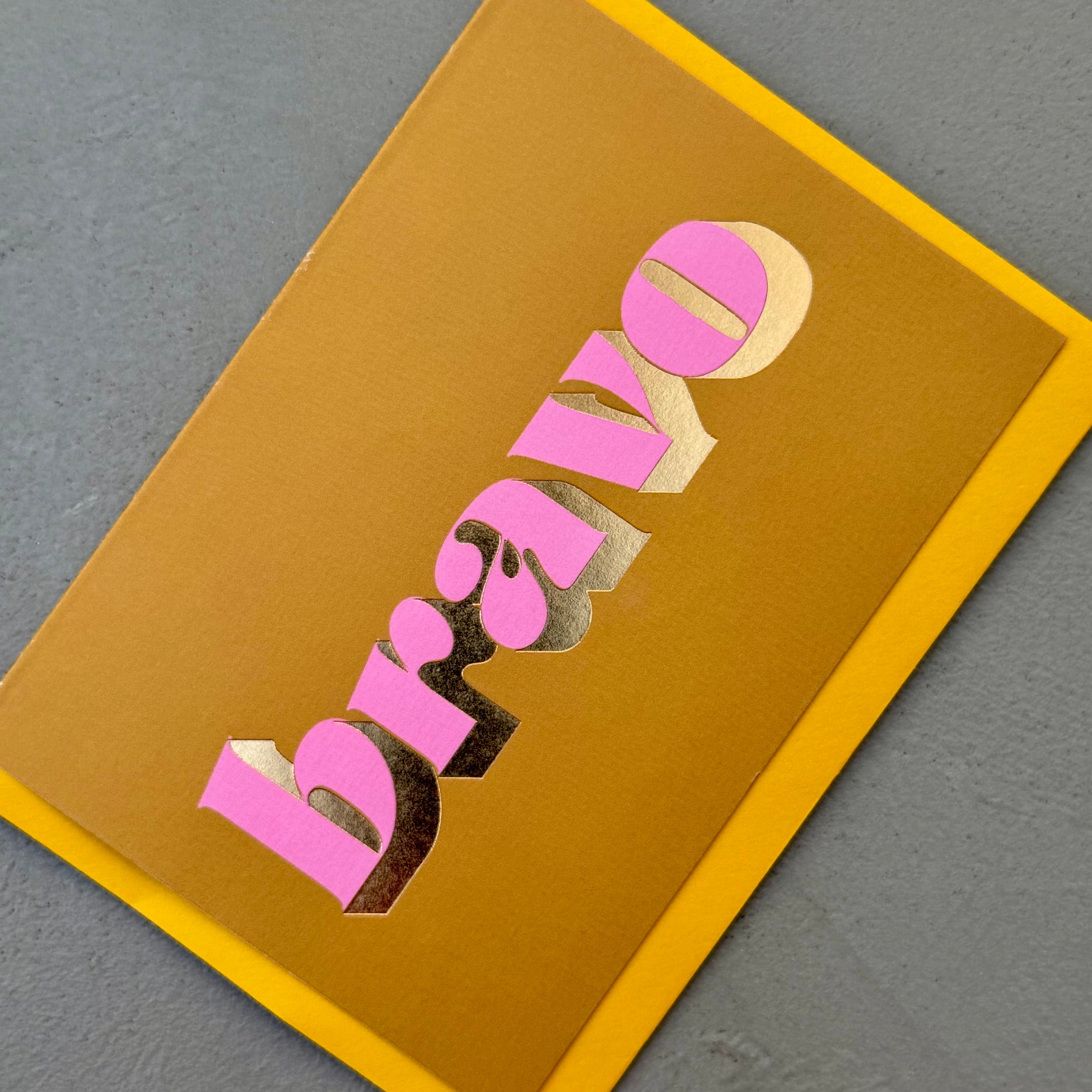Angled view of the ochre Bravo card showing the pink lettering and reflective gold foil.
