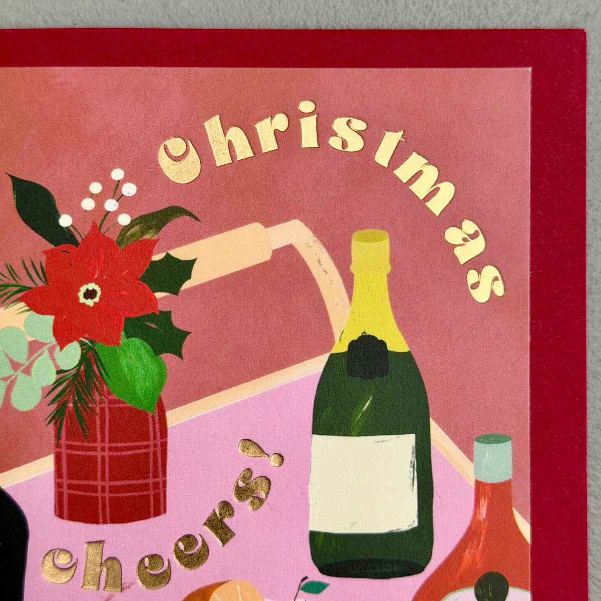 Close-up of Christmas Cheers card showing gold foil lettering, champagne bottle, flowers and festive drinks
