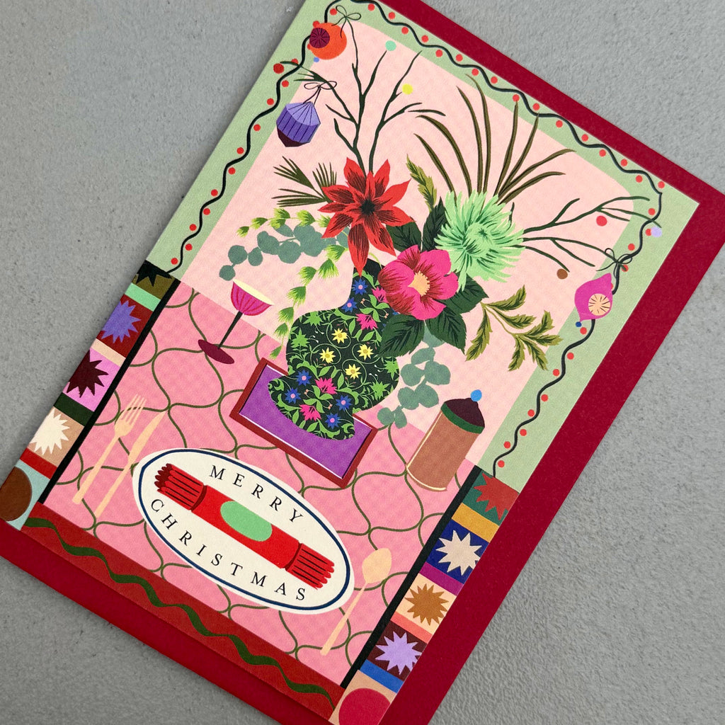 Angled view of Christmas Cracker card with illustrated festive tablescape and red envelope
