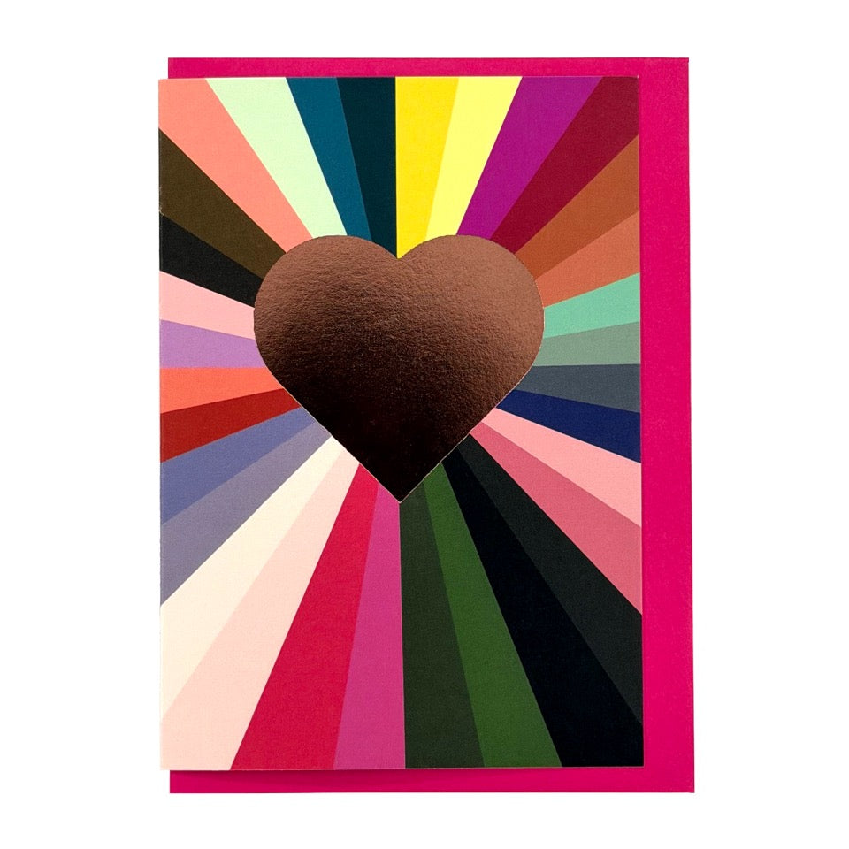 Greeting card featuring a gold foil heart with colourful radiating stripes on a bright pink envelope.
