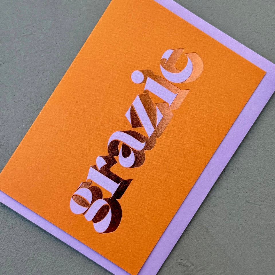 Angled view of the orange Grazie card showing the lilac lettering and reflective copper foil.