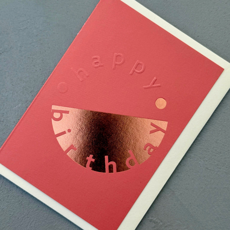 Angled view of the coral pink Happy Birthday card showing the embossed lettering and reflective copper foil.