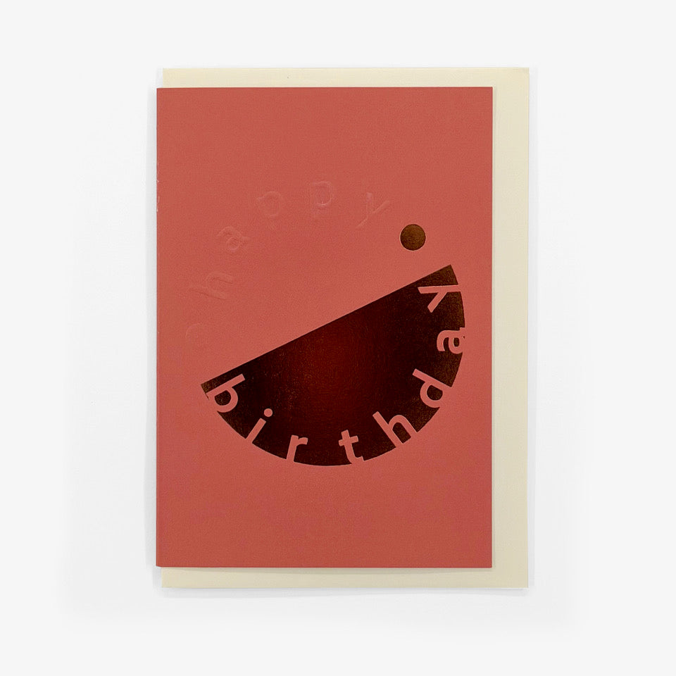 Coral pink Happy Birthday card with embossed lettering and copper foil graphic, shown with a cream envelope.
