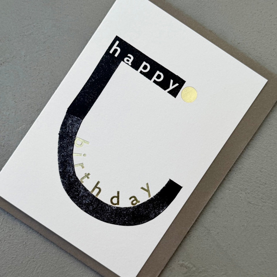 Angled view of the Happy Birthday Typography greeting card with a warm grey envelope, showcasing the minimalist black and white design with gold foil accents.
