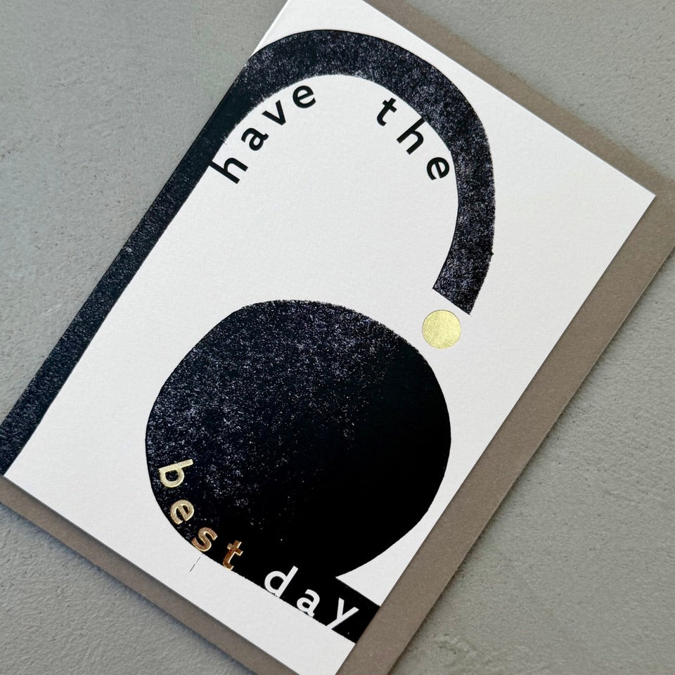 Angled view of the Have the Best Day greeting card with a warm grey envelope, showcasing the minimalist black and white design with gold foil details.