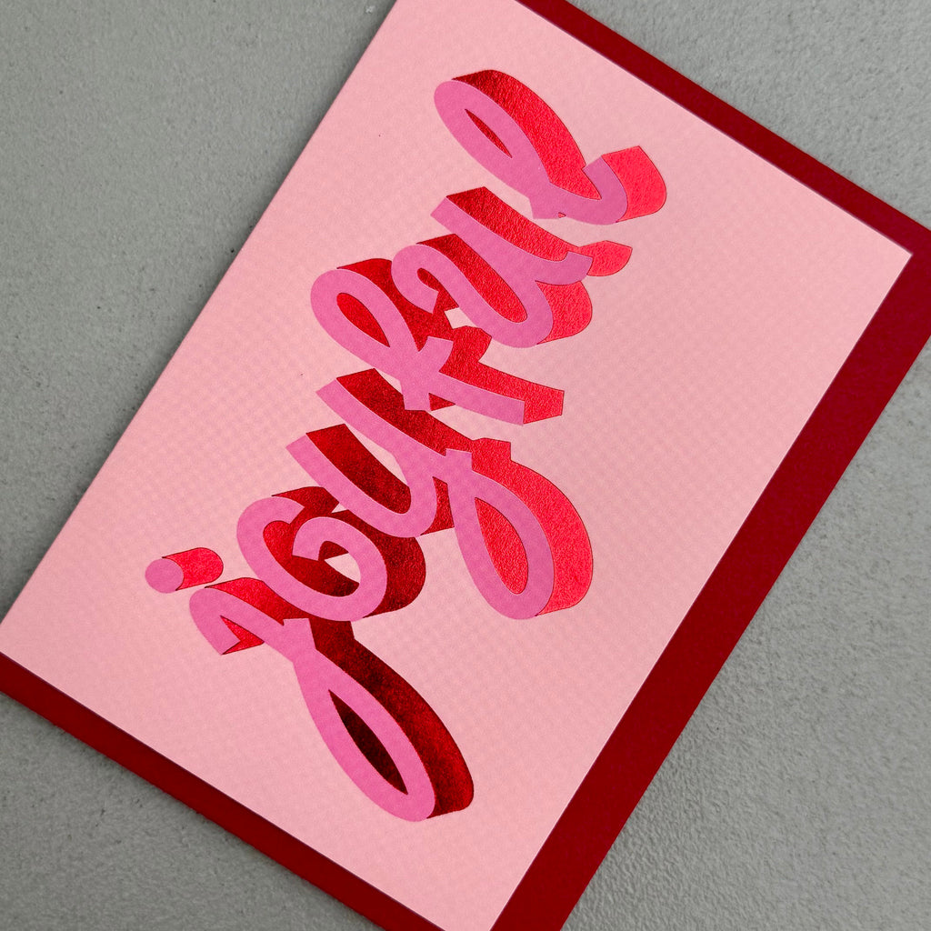 Angled view of Joyful Christmas card by PAVILION with sculptural pink lettering, red foil details and crimson envelope