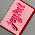 Angled view of Joyful Christmas card by PAVILION with sculptural pink lettering, red foil details and crimson envelope