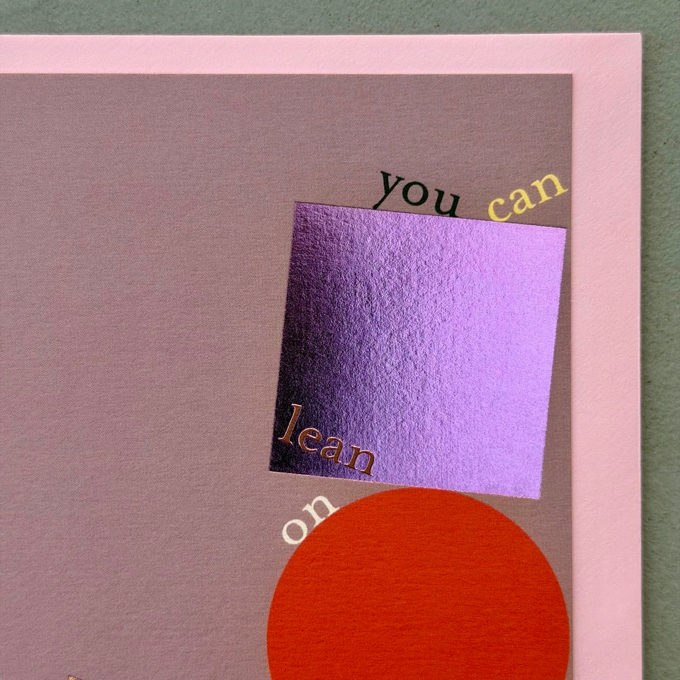 Close-up of the Lean On Me greeting card highlighting the metallic foil square and triangle, vibrant red circle and elegant typography on a muted mauve background.
