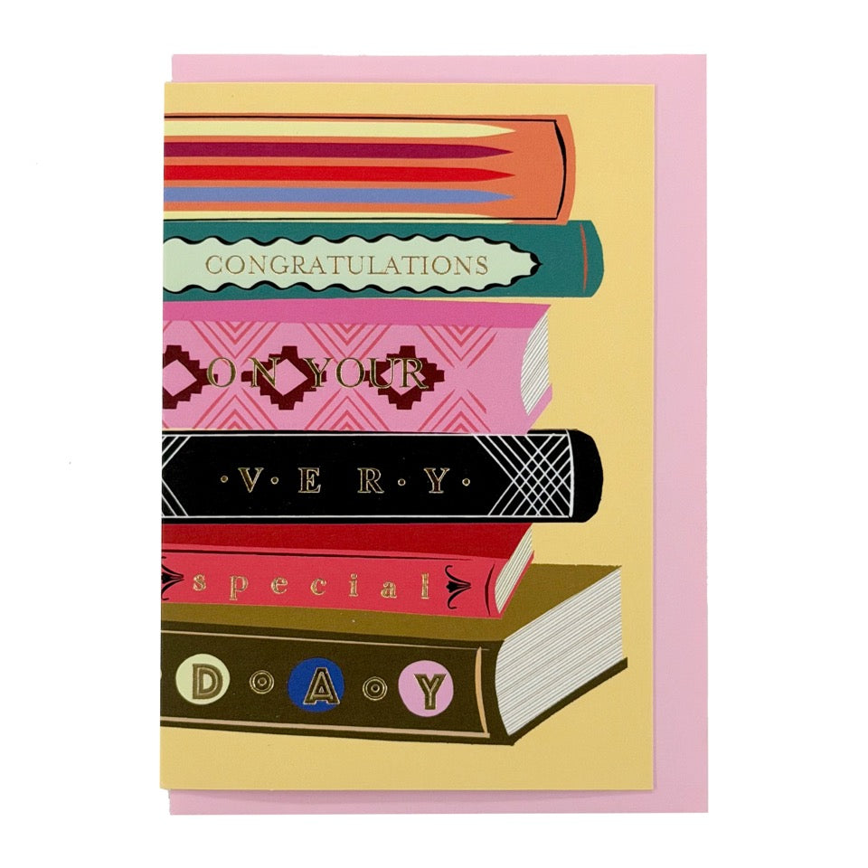 Congratulations card featuring a colourful stack of illustrated books, shown with a pale pink envelope.