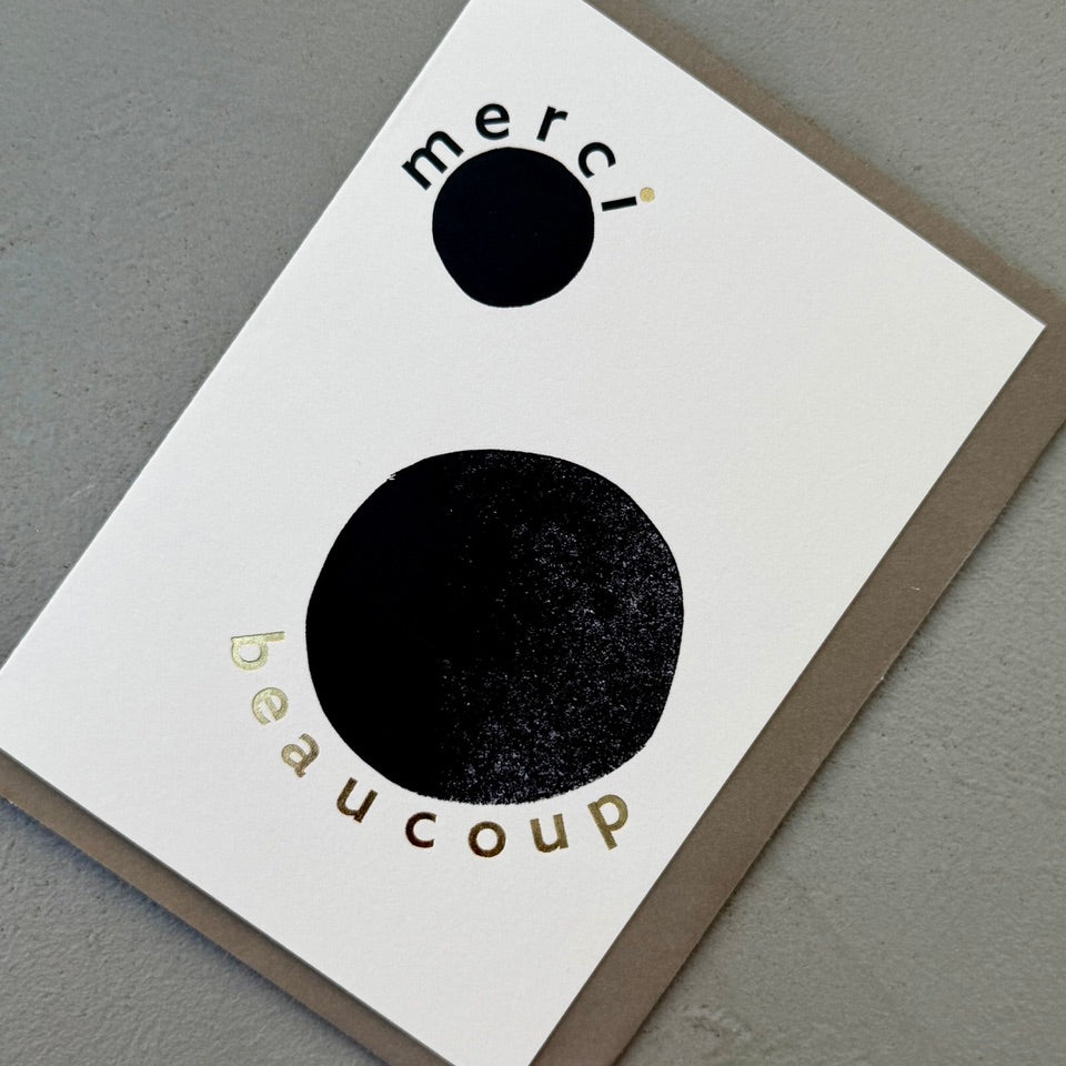 Angled view of the Merci Beaucoup greeting card with a warm grey envelope, showcasing the minimalist black and white artwork with gold foil details.