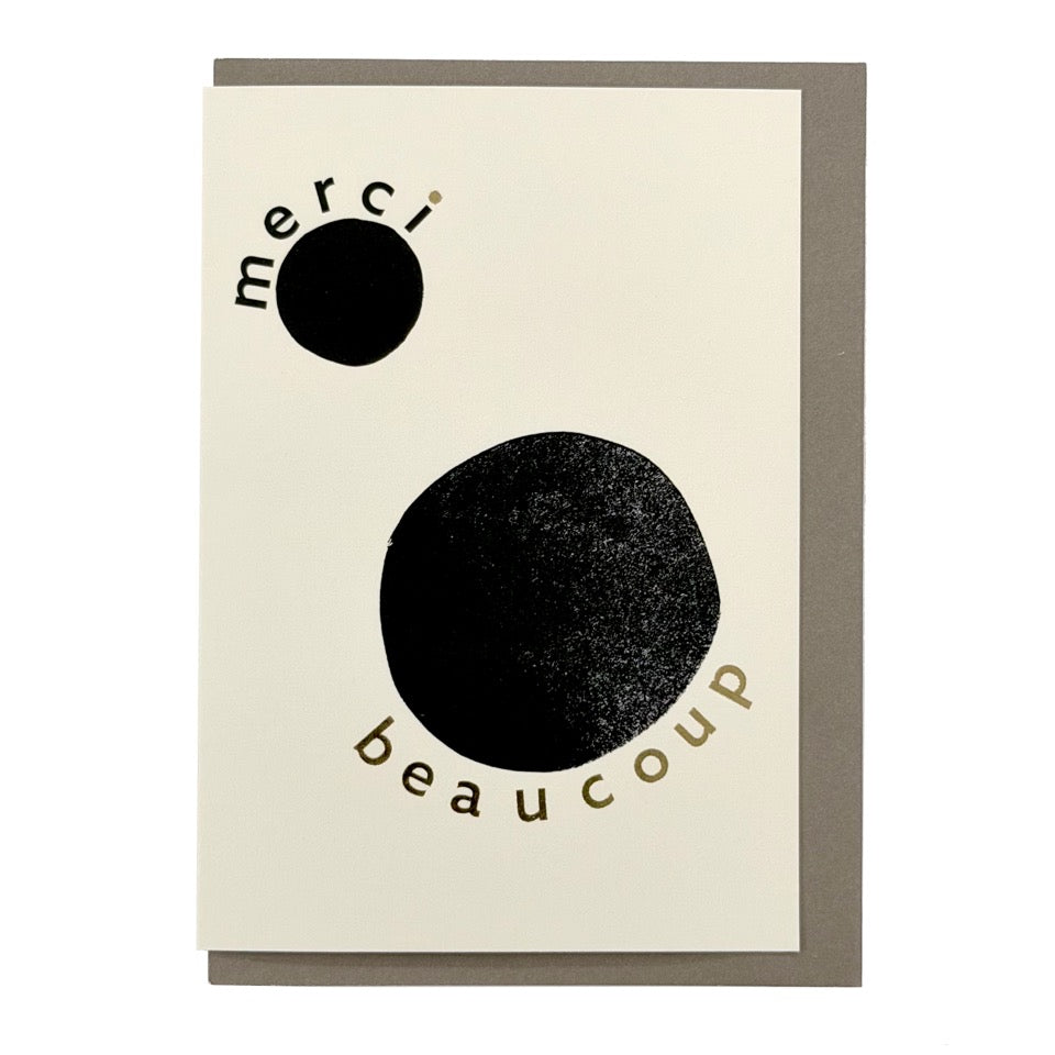 Minimalist thank you card featuring bold black abstract circles, gold foil typography reading "Merci Beaucoup", and a warm grey envelope.