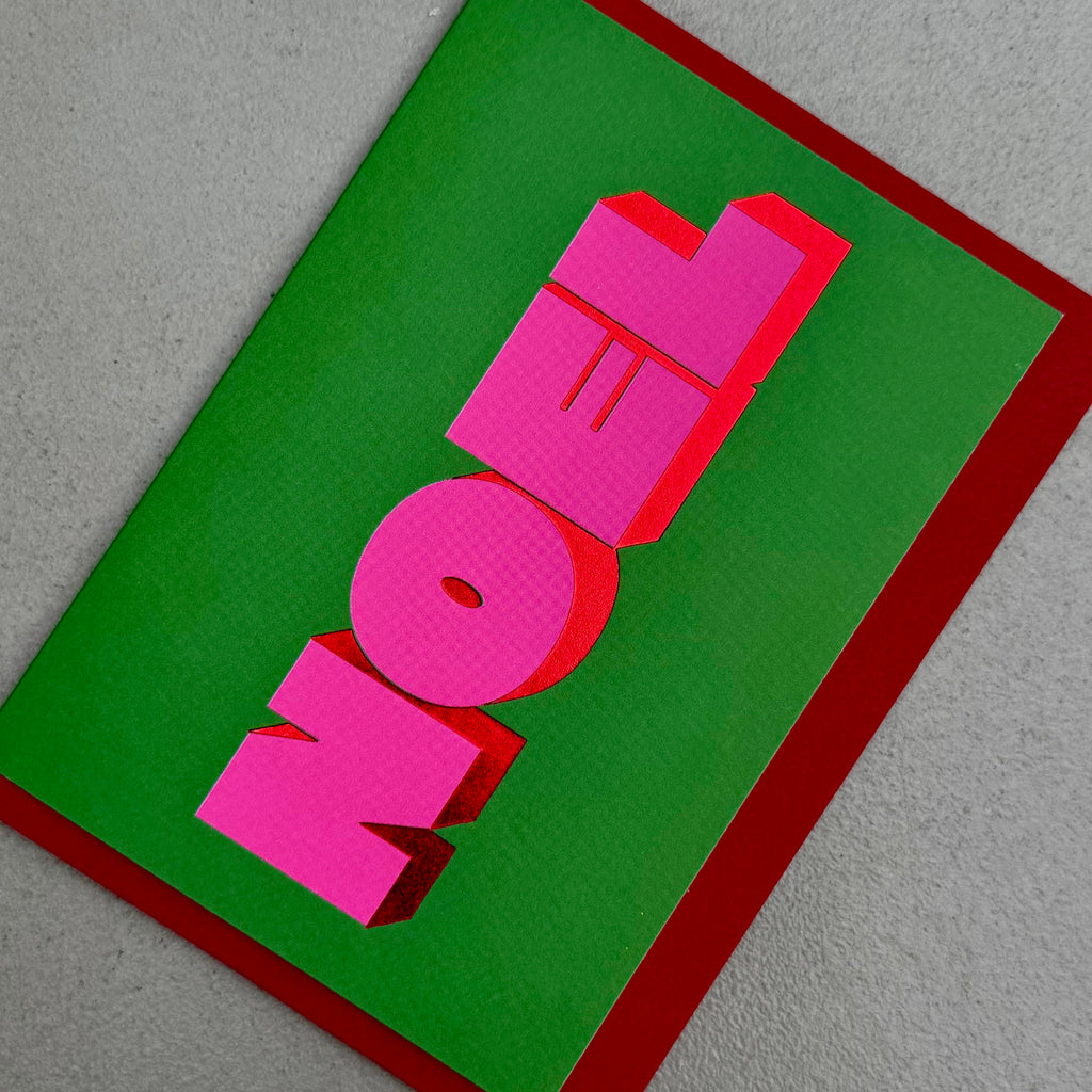 Angled view of Noel Christmas card by PAVILION with bold pink 3D lettering on a green background and red envelope