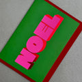 Angled view of Noel Christmas card by PAVILION with bold pink 3D lettering on a green background and red envelope