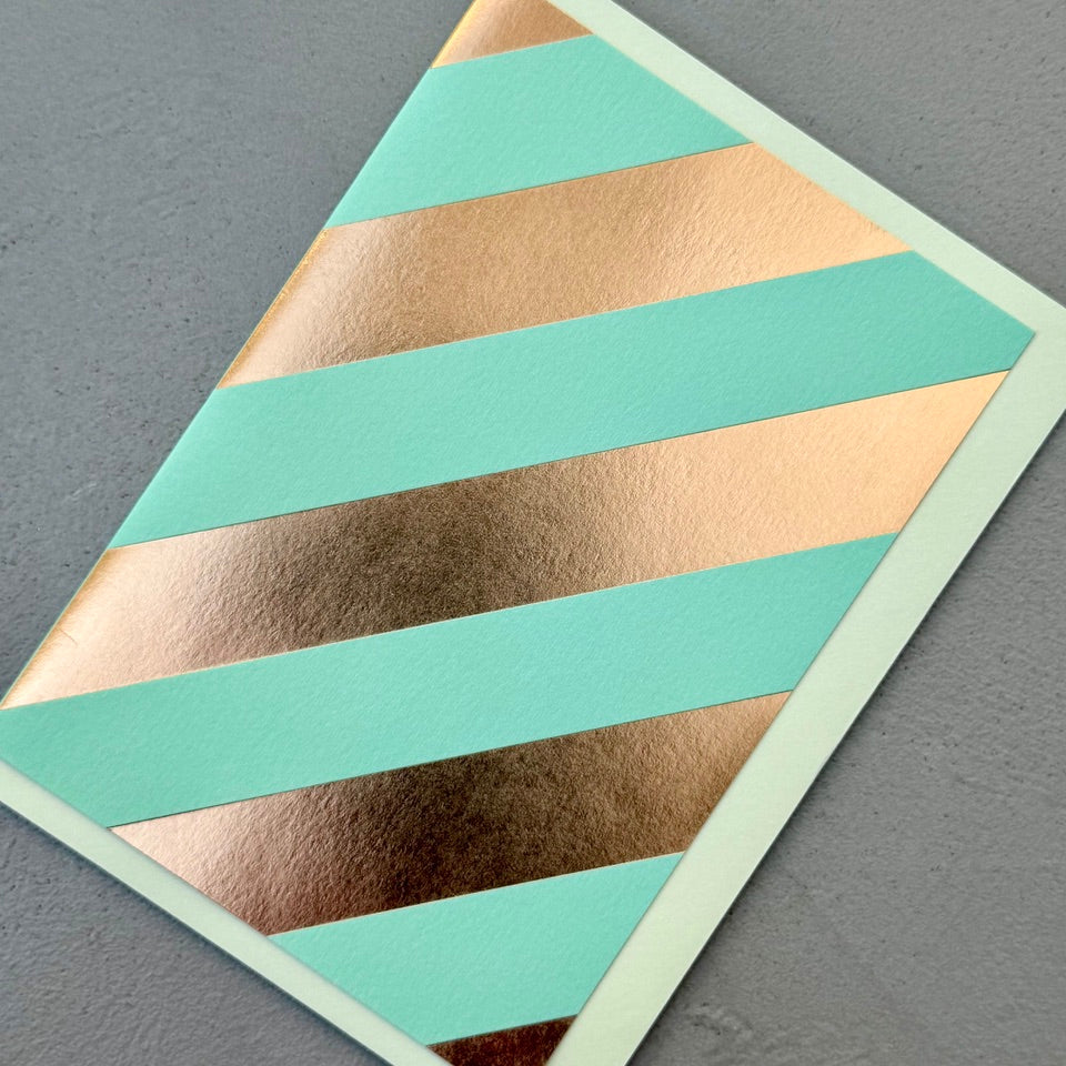 Angled view of the Oblique greeting card with a pale mint envelope, showcasing the bold diagonal copper foil pattern on a soft mint background.