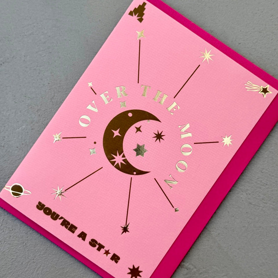 Angled view of the Over the Moon You're a Star greeting card with a bright pink envelope, showcasing the celestial artwork and shimmering foil finish.