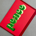 Angled view of Peace Christmas card by PAVILION with sculptural green lettering, gold foil details and red envelope