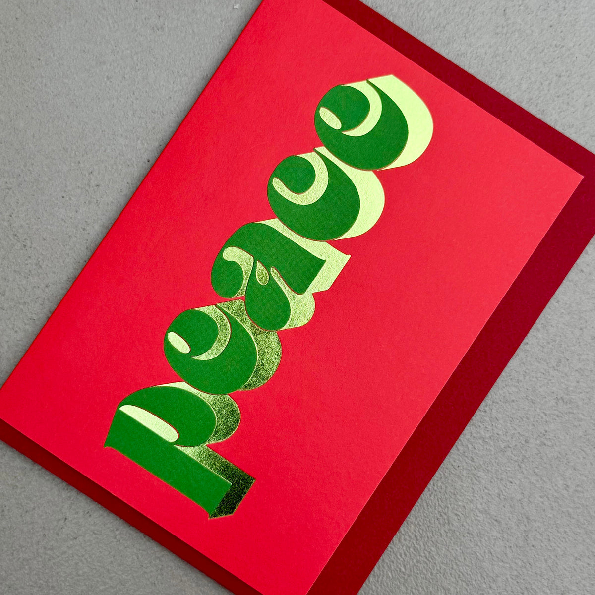 Angled view of Peace Christmas card by PAVILION with sculptural green lettering, gold foil details and red envelope