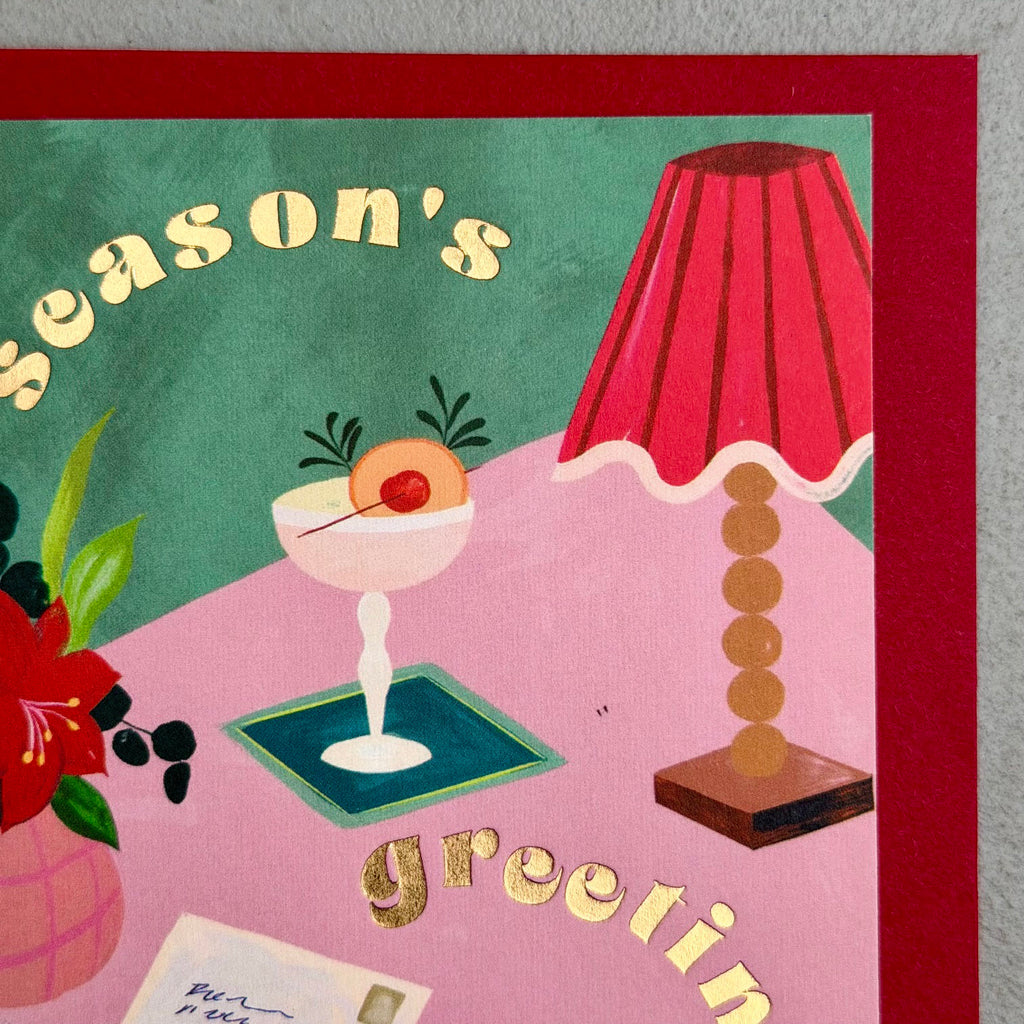 Close-up of Season's Greetings card showing gold foil lettering, cocktail, lamp and festive floral details