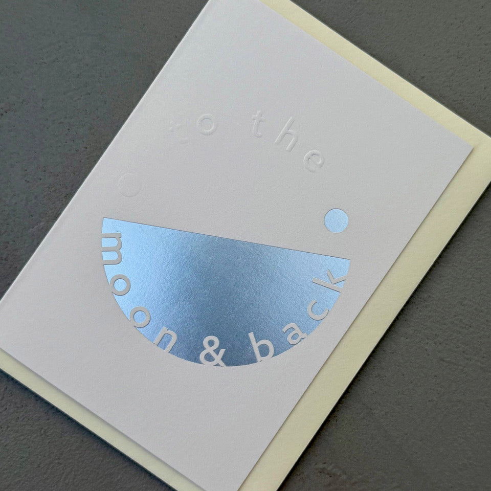Angled view of the pale grey To the Moon & Back card showing the embossed lettering and reflective silver foil.
