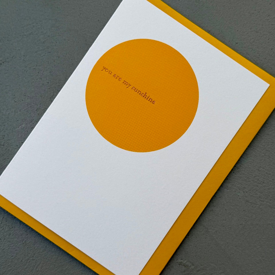Angled view of the You Are My Sunshine greeting card with a mustard yellow envelope, showcasing the clean minimalist design and elegant gold foil detail.