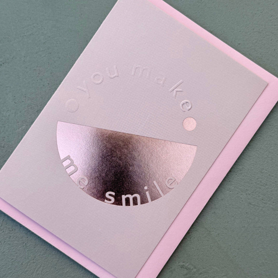 Angled view of the pale lilac You Make Me Smile card showing the embossed lettering and reflective pink foil.

