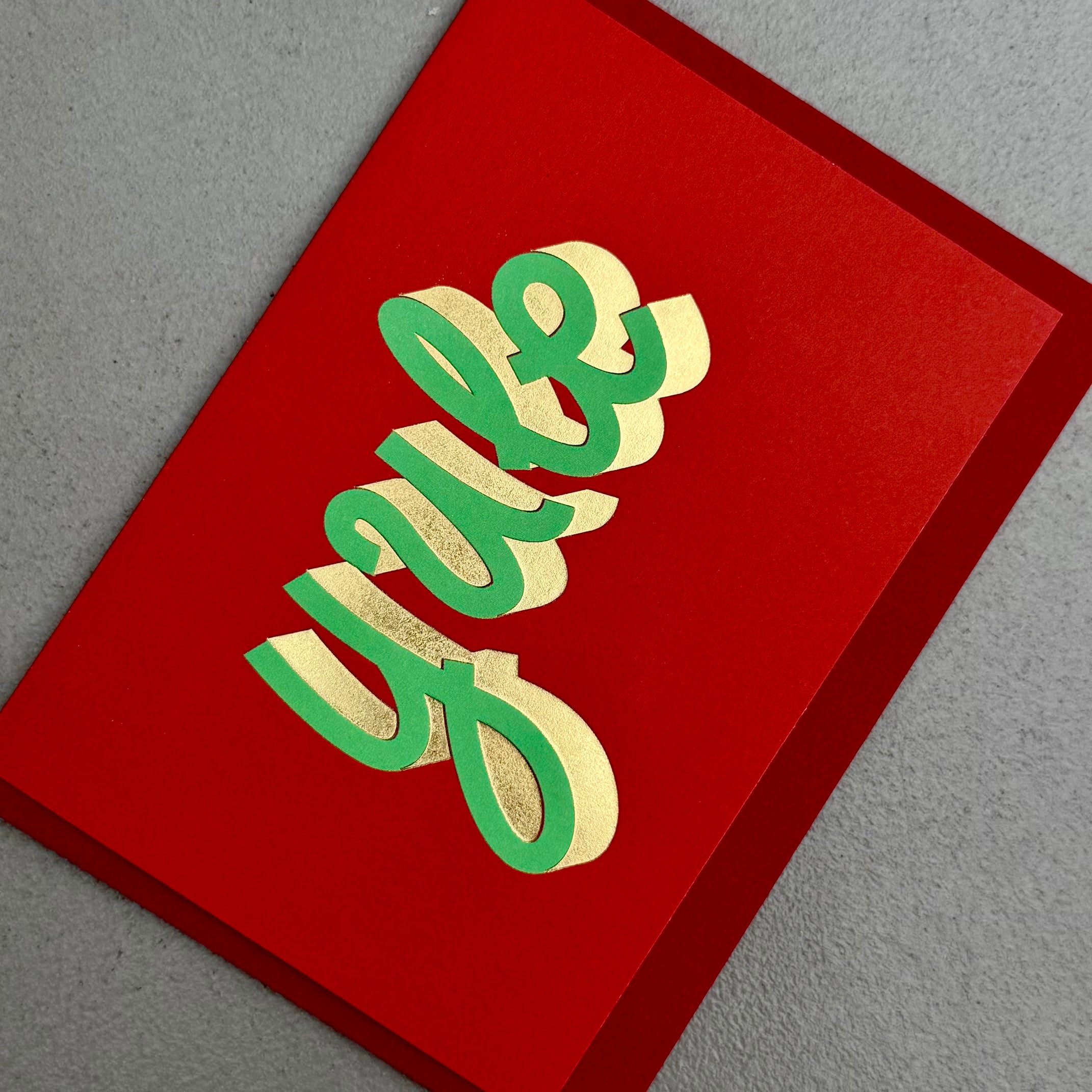 Angled view of YULE Christmas card by PAVILION with sculptural green lettering, gold foil details and red envelope